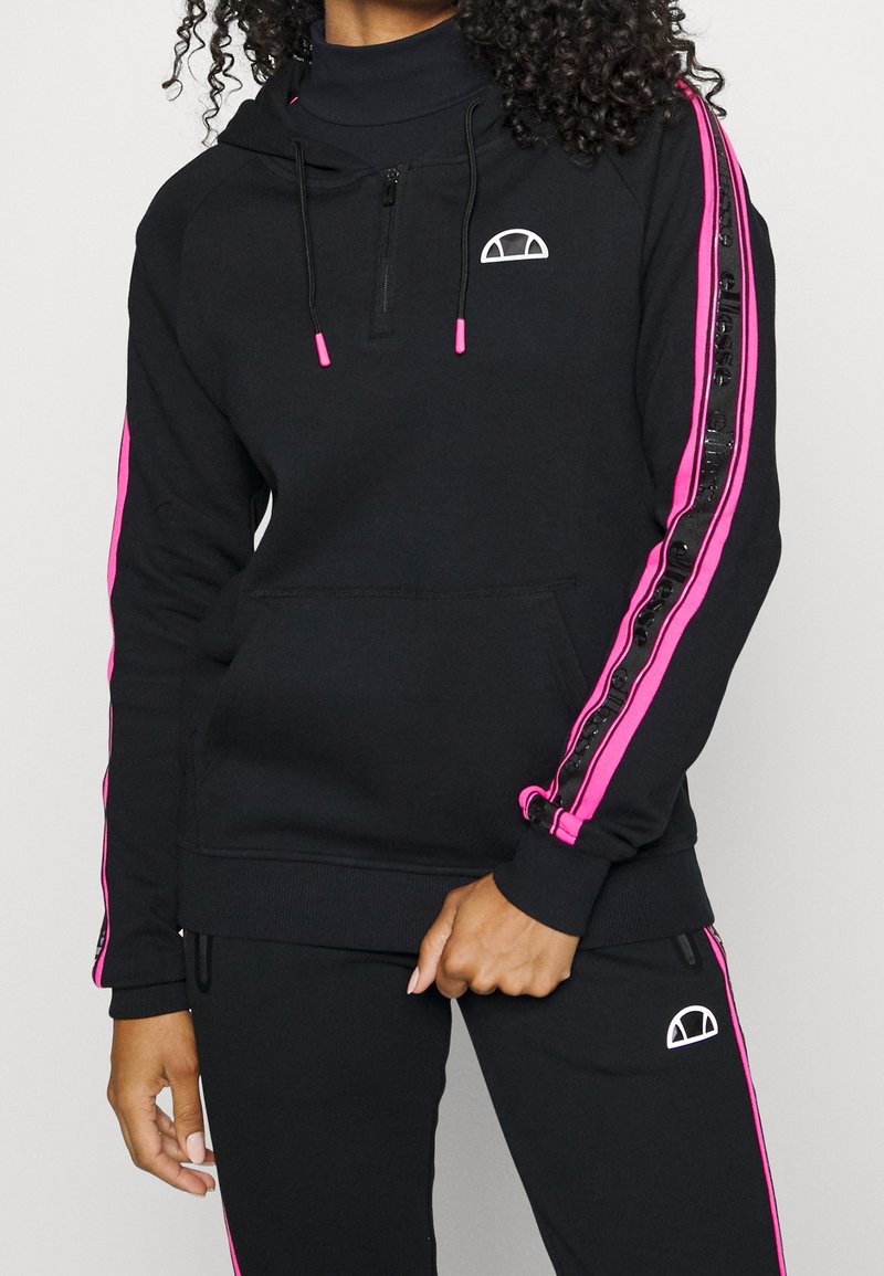 Femme portant un survêtement noir avec des bandes roses et des logos blancs, tenant une main près de la taille sur un fond clair uni.