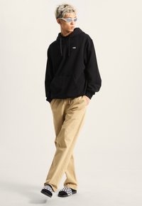 Felpa nera con tasca frontale e logo bianco, pantaloni beige a vestibilità larga e scarpe slip-on nere a quadretti. Il modello ha capelli corti e chiari e occhiali da sole.