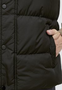 Schwarze gepolsterte Weste aus glänzendem Stoff, mit Stehkragen, Druckknöpfen und Taschen an den Seiten. Weicher Stoff mit gesteppten Abschnitten.