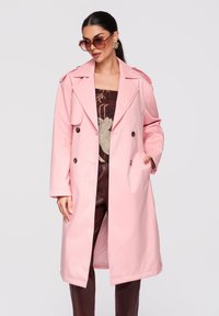 Femme portant un trench-coat rose oversize, de grandes lunettes de soleil, des boucles d'oreilles dorées, un haut à motifs et un pantalon bordeaux sur un fond uni.