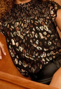 Black top with a smocked neckline featuring a leopard print in gold and bronze tones, lightweight material, short sleeves, and a loose fit.