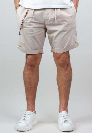 Pantaloni corti di cotone beige con risvolti, dotati di vita con coulisse e una tasca laterale con decorazioni in perline. Indossati con scarpe da ginnastica bianche.