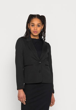 Blazer noir avec une coupe ajustée, revers crantés, deux poches avant et fermeture à un bouton. Porté par-dessus une robe noire côtelée.