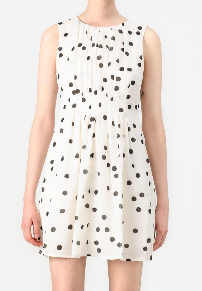 Femme portant une robe blanche sans manches à pois noirs, avec des plis sur le devant et une jupe évasée.