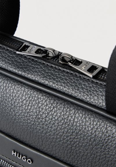 Gros plan sur un sac HUGO noir en cuir grainé, doté de deux tirettes de fermeture éclair en métal poli.