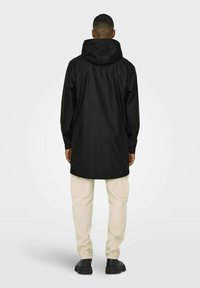 Jachetă neagră impermeabilă cu glugă, croi lejer, cu un tiv mai lung în spate, asortată cu pantaloni cargo bej deschis și pantofi negri.