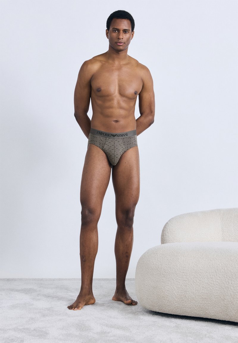 Καφέ ανδρικά εσώρουχα Emporio Armani με σχέδιο και λωρί�δα μέσης με λογότυπο, φορεμένα από μοντέλο που στέκεται σε ανοιχτό χαλί.
