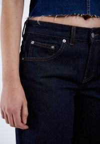 Jeans de mezclilla azul oscuro con costuras naranjas, cinco bolsillos y una cintura ajustada. La tela tiene una textura suave, mostrando un desgaste mínimo.