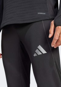 Schwarze Sportleggings mit glatter Textur, die ein auffälliges silbernes Adidas-Logo und ein atmungsaktives Aeroready-Label am Bund aufweisen.