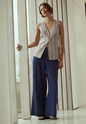 Kobieta stojąca wewnątrz, trzymająca pionowy drewniany panel, ubrana w beżową prążkowaną kamizelkę bez rękawów i szerokie granatowe spodnie z czarnymi sandałami.