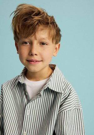 Jeune garçon aux cheveux châtain clair ébouriffés portant une chemise rayée verticale et un sous-vêtement blanc, sur un fond bleu clair.
