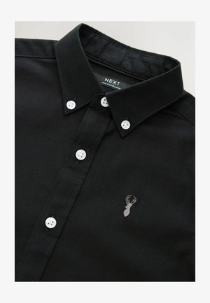 Juodas medvilninis marškinėlių švarkelis su apykakle, kuriame yra balti sagos ir mažas pilkas siuvinėtas elnio logotipas ant krūtinės.