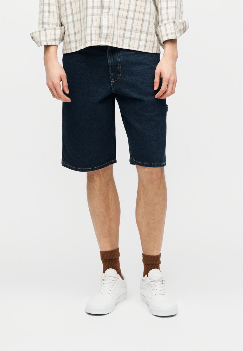 Uomo con shorts in denim scuro, sneaker bianche, calzini marroni e camicia a quadri a maniche lunghe arrotolata, in piedi su sfondo bianco.