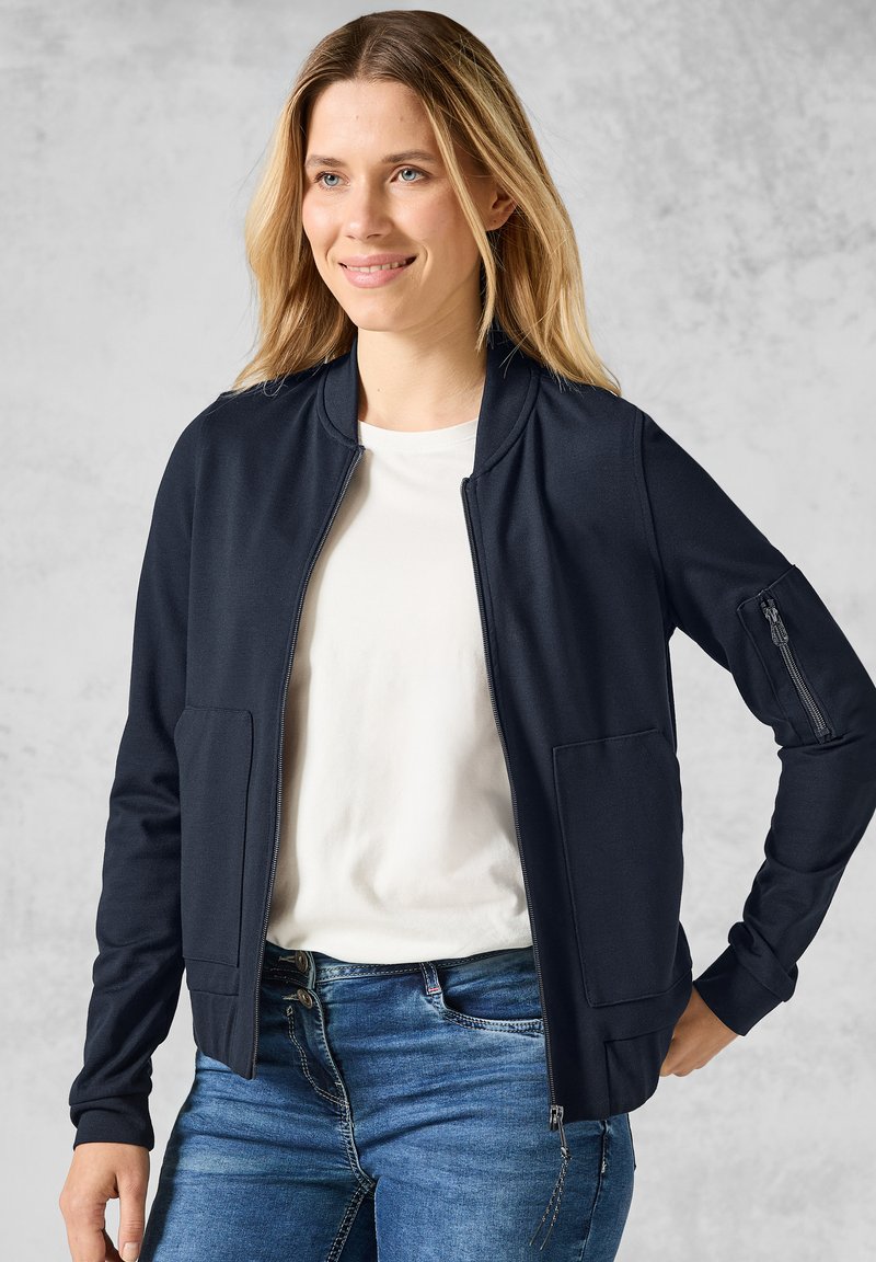 Giacca zip-up di un blu navy scuro con una leggera lucentezza, dotata di due tasche frontali e una tasca sulla manica, indossata sopra una t-shirt bianca e jeans blu.