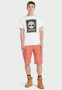 Jaunas vyras, vilkintis baltu Timberland logotipu pažymėtu marškinėliu, lašišinių spalvų cargo šortais, juodomis kojinėmis ir rusvais darbo batais, stovi prieš vienodą foną.