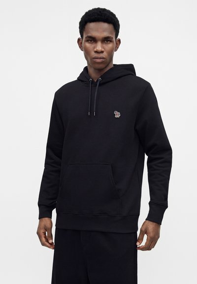 Homme portant un sweat à capuche noir avec un petit logo animal coloré sur la poitrine et une poche avant, debout devant un fond clair uni.