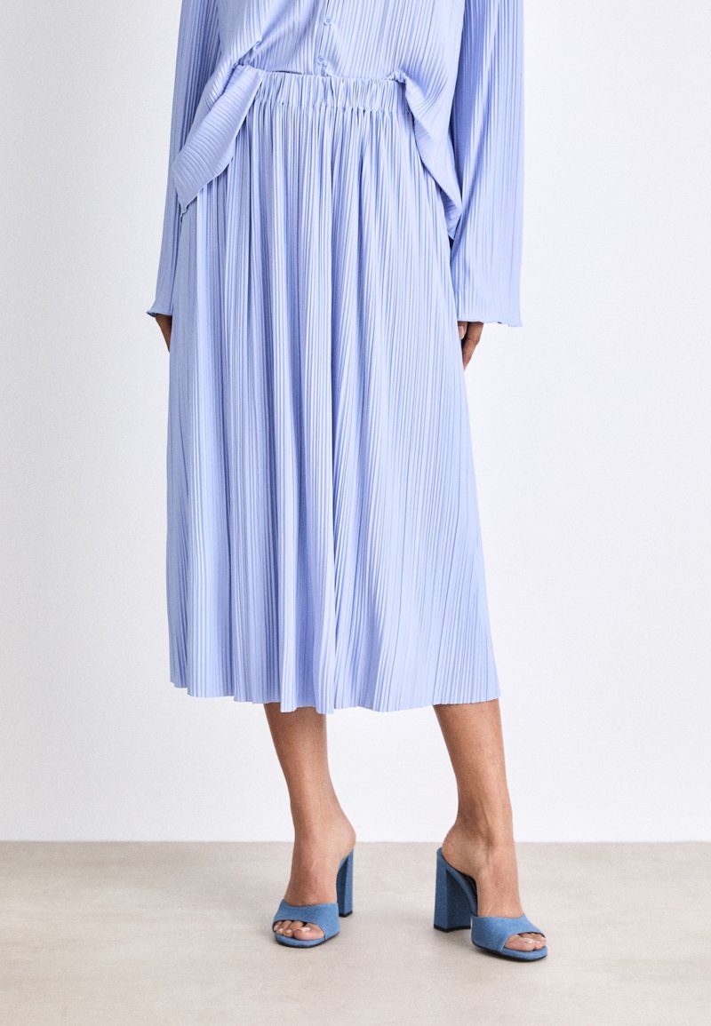 Femme portant une jupe midi plissée bleu clair, un haut assorti à manches longues et des sandales à talons carrés bleues à bout ouvert, debout sur un sol neutre.
