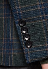 Skarpetkowy blezer wełniany w kratkę w ciemnozielonych, niebieskich i brązowych odcieniach. Cztery czarne guziki o gładkiej fakturze na mankiecie. Brak widocznych wzorów na materiale.