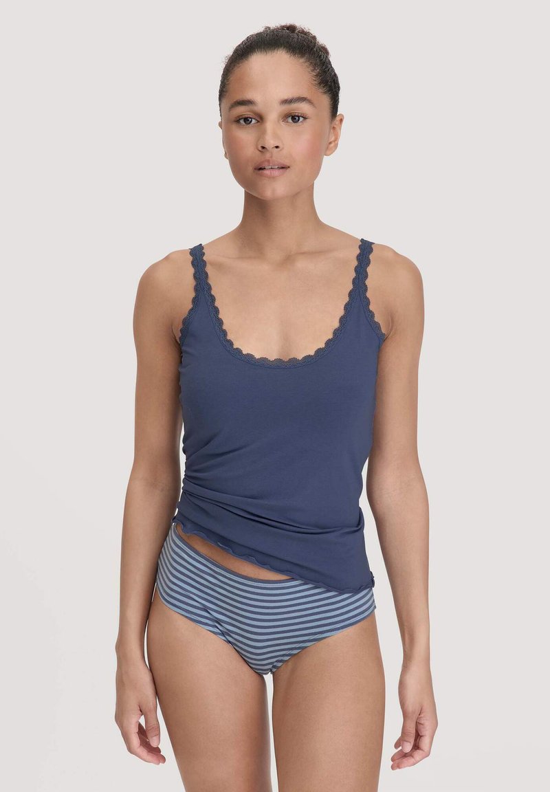 Vrouw in een marineblauwe tanktop met kant en blauwe gestreepte onderbroek, staand tegen een effen lichte achtergrond.