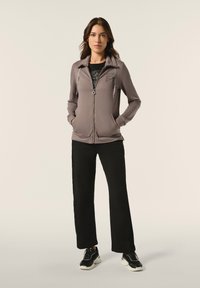 Giacca zip grigia chiara con colletto, tasche frontali e logo. Abbinata a pantaloni neri a gamba larga e scarpe sportive nere.