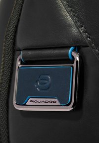 Zwart leren product met een rechthoekig blauw metalen accent, voorzien van een zilveren logo en stikdetails, tegen een gestructureerde achtergrond.