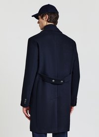 Cappotto in lana blu navy con vestibilità sartoriale, ampi revers e dettaglio cintura posteriore. Presenta polsini con bottoni e una texture liscia.