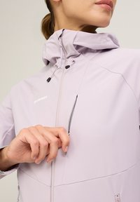 Ljus lavendelfärgad, vattentät softshell-jacka med hög krage, dragkded ficka och justerbar huva. Har en slät textur och atletisk passform.