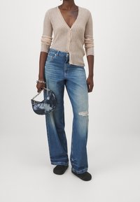 Cardigan beige côtelé avec col en V et fermeture à boutons, associé à un jean bleu clair à jambes larges avec des détails usés. Porte un sac à main bleu tie-dye.