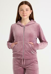 Mladý člověk s dlouhými hnědými vlasy, oblečený v mauve zipové sametové bundě s kapucí a odpovídajících kalhotách, s rukama v kapsách bundy.