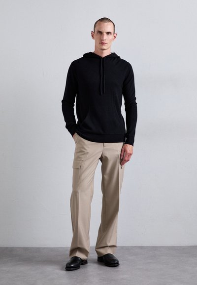 Svart hettegenser laget av myk stoff, parret med lys beige cargo-bukser med lommer, og svarte slip-on sko.