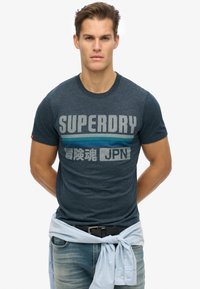 Camiseta de manga corta azul marino con estampado gráfico en gris y verde azulado. Presenta un cuello redondo y un corte relajado. El texto dice "SUPERDRY JPN."