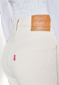 Jeans de mezclilla blanca con un diseño clásico, que presentan una etiqueta de cuero con los detalles de la marca y una pequeña etiqueta roja de la marca en el bolsillo trasero.