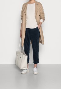Frau trägt einen beigen Trenchcoat, einen weißen Pullover, eine marineblaue Cordhose, weiße Turnschuhe und hält eine große weiße Handtasche, während sie vor einem weißen Hintergrund steht.