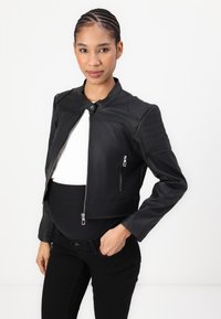 Chaqueta de cuero negra corta con cremalleras plateadas, detalles acanalados en los hombros y un cuello con broche, llevada sobre una blusa blanca ajustada y pantalones negros.