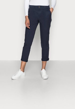 Femme portant un pantalon court bleu marine retroussé, un haut blanc à manches longues et des baskets blanches, debout avec une main dans la poche.