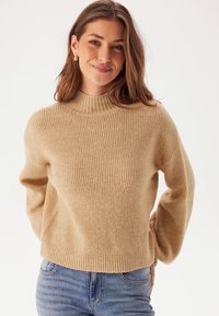 Beige gestrickter Pullover mit hohem Rundhalsausschnitt, gerippter Textur und Ballonärmeln; über Jeans aus Denim in einem neutralen Hintergrund getragen.