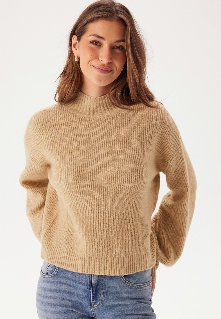 Beige gestrickter Pullover mit hohem Rundhalsausschnitt, gerippter Textur und Ballonärmeln; über Jeans aus Denim in einem neutralen Hintergrund getragen.