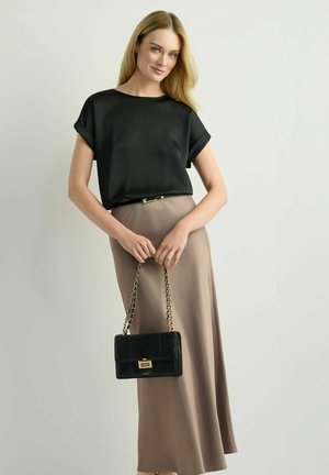 Kobieta ubrana w czarną bluzkę z krótkim rękawem i długą spódnicę koloru taupe, trzymająca czarną torebkę na złotym łańcuszku na jednolitym tle.