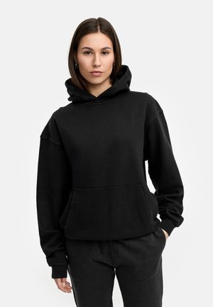 Schwarzer Kapuzenpullover mit einer Vordertasche, lockerer Passform, weichem Material und gerippten Bündchen. Kombiniert mit passenden schwarzen Hosen.