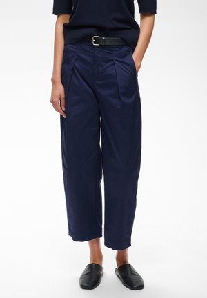 Pantalon large cropped bleu marine avec plis, ceinture noire, chaussures noires à enfiler et haut bleu marine à manches courtes sur une personne debout.