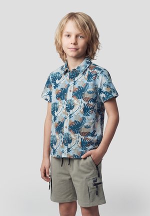 Niño rubio con cabello de longitud media, vistiendo una camisa de manga corta azul con estampado tropical y pantalones cortos cargo beige, de pie con una mano en el bolsillo.