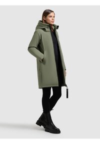 Cappotto verde oliva con cappuccio, dotato di zip frontale, tasche laterali e finitura texturizzata. Leggings neri e stivali neri chunky completano l'outfit.