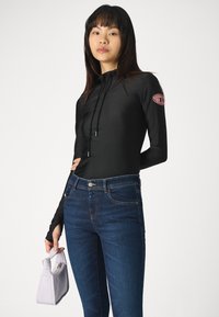 Μαύρη μπλούζα με μακριά μανίκια και κουκούλα, διακοσμημένη με ροζ λογότυπο στον αριστερό βραχίονα, σε συνδυασμό με σκούρα μπλε τζιν και μια μικρή λευκή τσάντα.