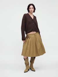 Femme portant un cardigan boutonné marron foncé, une jupe midi plissée couleur fauve et des bottes amples assorties couleur fauve, debout devant un fond clair uni.
