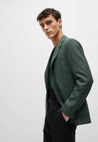 Blazer verde con texture dal taglio su misura, design monopetto, rever a notches e tasche laterali, abbinato a un top e pantaloni neri.