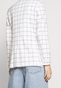 Blazer blanc avec un motif à carreaux en lignes noires, doté d'épaules structurées et d'une fente au dos. Assorti à un pantalon en denim bleu clair.