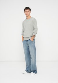 Suéter de malha canelada cinza claro com gola redonda, combinado com jeans de denim azul claro de corte solto e ténis brancos, de pé contra um fundo plain.