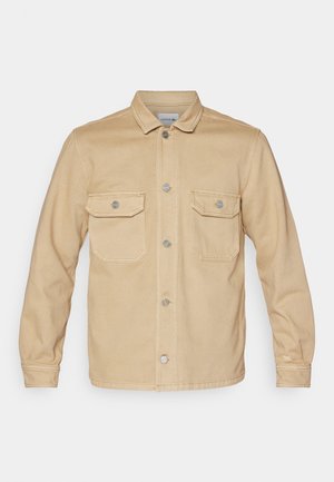 Chaqueta de mezclilla beige con cierre de botones en la parte delantera, dos bolsillos en el pecho y un cuello clásico. Presenta detalles de costura y botones de hardware plateado.