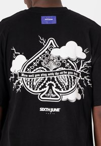 Camiseta de algodón negra con un gran gráfico blanco de un diseño de pica ornamentado con nubes, rayos y el texto "Qué bien juegas con las cartas que tienes en la mano."