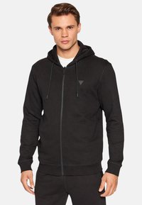 Schwarzer Kapuzenpullover mit durchgehendem Reißverschluss, Kordelzugkapuze, gerippten Bündchen und einem kontrastierenden dreieckigen Logo auf der Brust. Hergestellt aus weichem Stoff.